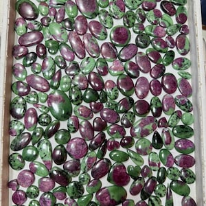 May include: A tray filled with ruby zoisite gemstones in a variety of oval and rounded forms. The stones are a mix of deep green and rich magenta, with a polished surface. The gemstones are displayed in a rectangular tray.