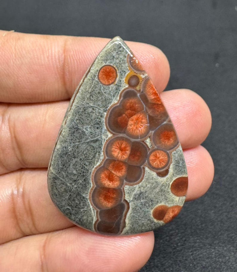 May include: A teardrop-shaped gemstone with a unique pattern. The stone features a gray base with intricate veining and is adorned with clusters of reddish-orange, flower-like formations. The polished surface highlights the natural colors and textures of the stone.