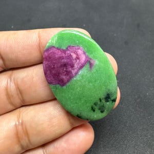 May include: Oval-shaped gemstone with a vibrant green base and a prominent, heart-shaped patch of deep magenta. The stone has a polished surface and small black inclusions.