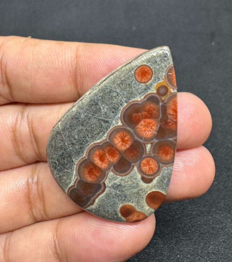 May include: A polished, teardrop-shaped gemstone with a gray base and intricate patterns of reddish-orange and brown circular designs. The stone is held in a hand, showcasing its unique color and texture. The gemstone is a natural, decorative item.