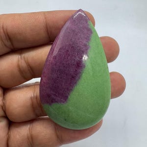 May include: A polished, teardrop-shaped gemstone. The stone is split diagonally, with a deep purple section on the left and a light green section on the right. The gemstone is held in a hand.