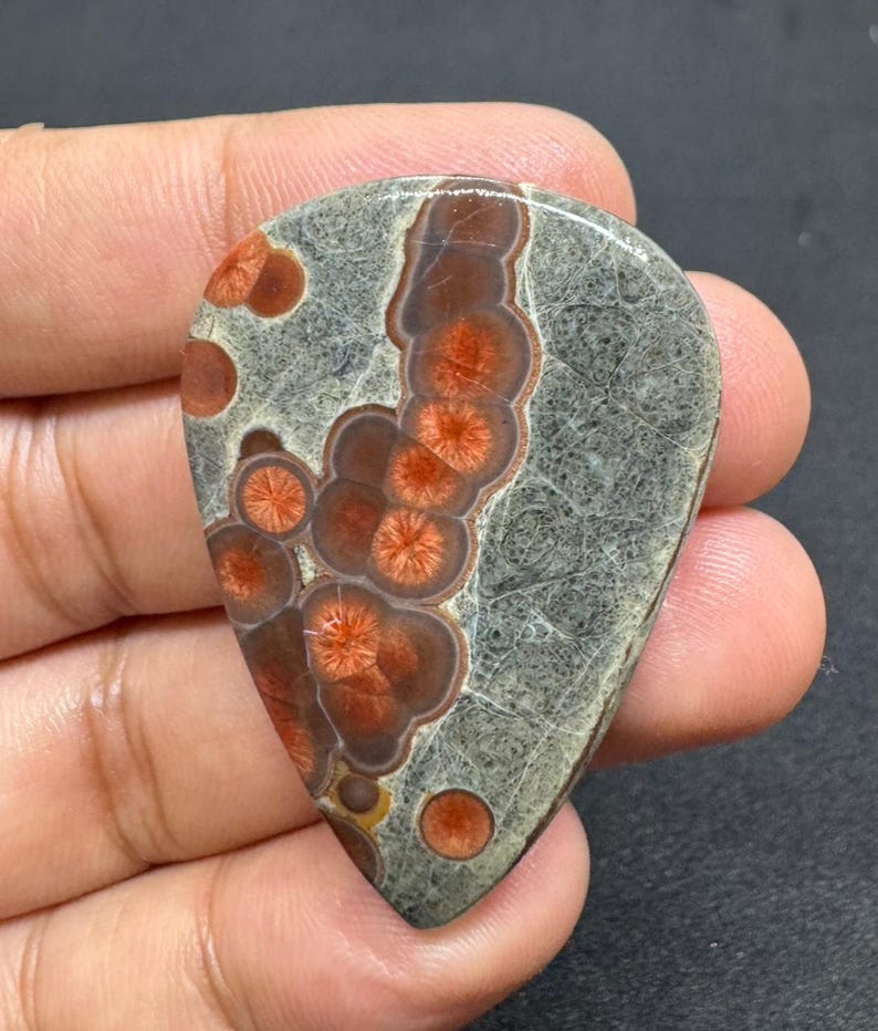 May include: A teardrop-shaped gemstone with a gray base and intricate patterns of reddish-brown and orange circular designs. The stone has a polished surface and is held in a hand, showcasing its unique color and texture.