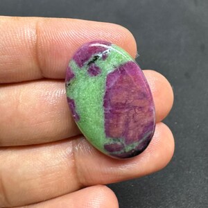 May include: Oval-shaped gemstone featuring a vibrant mix of colors. The stone showcases a striking combination of deep magenta and light green hues, with a polished, glossy surface. The gemstone is held in a hand, highlighting its size and shape.