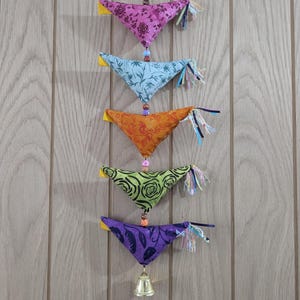 May include: A decorative mobile with five fabric triangles in various colours, including purple, blue, orange, green, and purple. Each triangle has a floral or botanical pattern. The mobile is decorated with beads, tassels, and a small gold bell at the bottom.