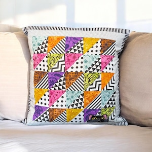 May include: Square cushion with a patchwork design. It features colourful triangles with patterns like stripes, polka dots, and floral prints. The cushion has a black and white striped border and a small black cat peeking out from the corner.