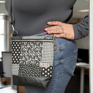May include: A black and white patchwork crossbody bag with a black leather bottom. The bag features various patterns, including polka dots, floral, and gingham. It has a silver zipper and a small tag with a dog illustration.
