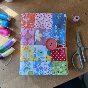Könnte beinhalten: Ein handgefertigtes Patchwork-Notizbuch mit einer Hülle aus Stoffquadraten in verschiedenen floralen und geometrischen Mustern. Die Farben umfassen Orange, Rosa, Blau und Grün. Ein rosa Knopf und ein Gummiband sichern das Notizbuch. In der Nähe befinden sich Garnrollen und eine Schere.