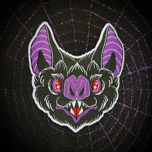 May include: A black and purple embroidered patch of a bat with a menacing expression. The bat has large, pointed ears and sharp teeth. The patch is perfect for adding a spooky touch to any project.