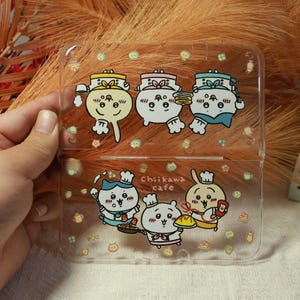 May include: Clear plastic case featuring cartoon characters in chef hats. The case has a hinged design and is decorated with food-related icons and the text "Chiiikawa cafe". The characters are in various poses.