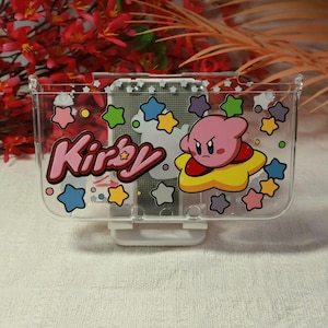 May include: Transparent plastic organiser with the word "Kirby" in pink and yellow, and a cartoon character. The design includes colourful stars and clouds. The organiser has a white base.