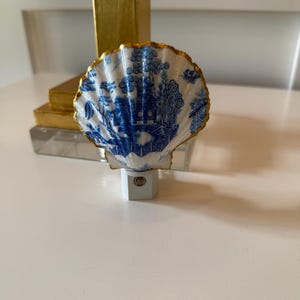 May include: A decorative night light featuring a seashell design. The shell is white with a blue Chinoiserie pattern and a gold-painted edge. The night light plugs into a standard wall outlet.