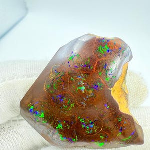Puede incluir: Una piedra de ópalo en bruto con un color base marrón oscuro. La piedra presenta destellos vibrantes de verde, azul y morado en toda su superficie. El patrón único y las variaciones de color del ópalo lo convierten en una gema llamativa.