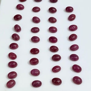 Puede incluir: Colección de gemas ovaladas de color rojo intenso dispuestas sobre una superficie blanca. Las gemas pulidas, posiblemente rubíes, tienen un aspecto liso y una saturación de color variable. La disposición sugiere gemas sueltas, aptas para joyería.