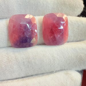 Puede incluir: Dos gemas facetadas, de talla cojín, con tonos rosados y púrpuras. Las gemas se muestran sobre un fondo de tela blanca, mostrando sus superficies pulidas y variaciones de color internas. Ideales para la creación de joyas.