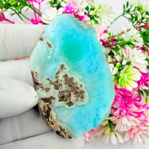 May include: A polished, irregular slab of bright turquoise blue gemstone featuring a central cluster of earthy brown and tan mineral inclusions. The stone has a smooth, translucent surface with natural color variations and a jagged, raw edge.