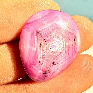 Puede incluir: Gema rosa ovalada y pulida, con un patrón en forma de estrella. La piedra, sostenida en una mano, muestra una estructura interna translúcida. El color rosa varía de claro a oscuro. La superficie pulida resalta los detalles.