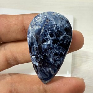 May include: A polished, teardrop-shaped gemstone with a swirling pattern of deep blue and white. The stone has a smooth, glossy surface and is held between two fingers. The overall appearance is of a natural, high-quality mineral.