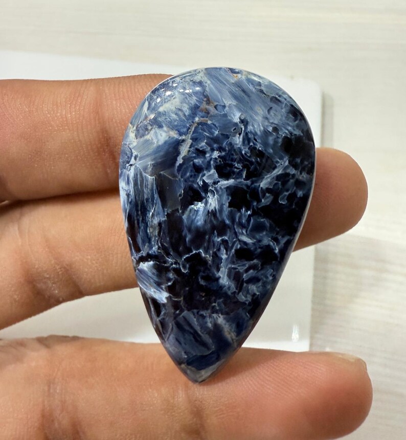 May include: A polished, teardrop-shaped gemstone with a swirling pattern of deep blue and white. The stone has a smooth, glossy surface and is held between two fingers. The overall appearance is of a natural, high-quality mineral.