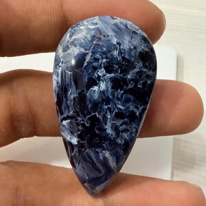 May include: A polished, teardrop-shaped gemstone with a striking pattern of deep navy blue and white swirls. The stone's surface is smooth and reflective, showcasing a natural, marbled appearance. The gemstone is held between fingers, highlighting its size and shape.