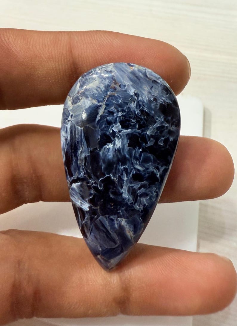May include: A polished, teardrop-shaped gemstone with a striking pattern of deep navy blue and white swirls. The stone's surface is smooth and reflective, showcasing a natural, marbled appearance. The gemstone is held between fingers, highlighting its size and shape.