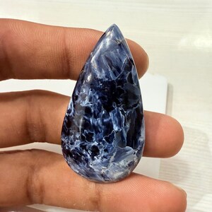 May include: A polished, teardrop-shaped gemstone with a striking blue and white pattern. The stone's surface has a marbled appearance, with swirling veins of white against a deep blue background. The gem is held between fingers.