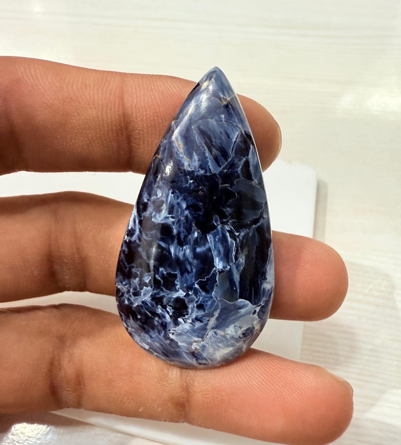 May include: A polished, teardrop-shaped gemstone with a striking blue and white pattern. The stone's surface has a marbled appearance, with swirling veins of white against a deep blue background. The gem is held between fingers.