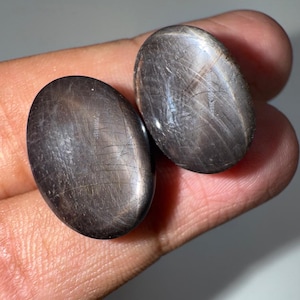 May include: Two oval-shaped, polished black star diopside gemstones. Each stone features a smooth, rounded surface with a visible chatoyant effect, displaying a subtle, light-reflecting line across the dark, charcoal-colored mineral structure.