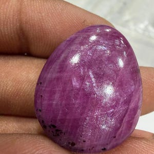 May include: A polished, oval-shaped gemstone in a deep magenta hue. The surface reflects light, showcasing internal textures and subtle color variations. Small, dark inclusions are visible throughout the stone. The gem is held in a hand, suggesting its size and form.