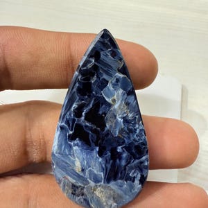 May include: A teardrop-shaped gemstone with a striking pattern of deep blue and lighter blue hues, with some areas appearing almost black. The stone has a polished surface and is held in a hand.
