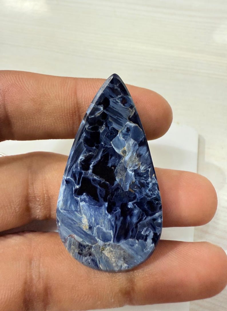 May include: A teardrop-shaped gemstone with a striking pattern of deep blue and lighter blue hues, with some areas appearing almost black. The stone has a polished surface and is held in a hand.