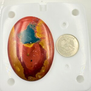 Puede incluir: Una gema ovalada pulida, que muestra una mezcla de tonos rojos, amarillos y azules. Una moneda con el texto "ПЯТЬ РУБЛЕЙ" y el año 2014 se encuentra junto a la gema, sirviendo como referencia de tamaño. La gema está sobre una superficie blanca.