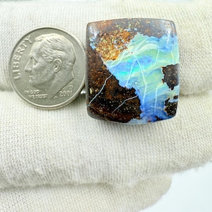 May include: A rectangular gemstone with a brown matrix and vibrant blue and green opal flashes. A British coin is next to the gemstone for size comparison. The opal has a unique pattern with cracks.