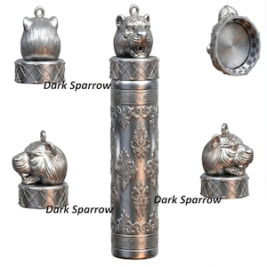 May include: A silver-coloured container with a detailed tiger head lid. The container has an ornate, floral pattern and the text "Dark Sparrow". The lid has a loop for hanging. The container is likely used for storing small items.