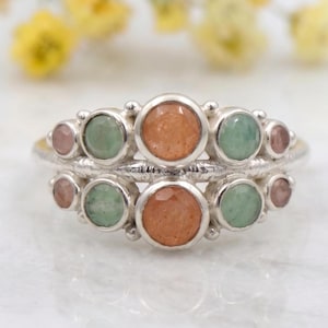 May include: Two silver rings with a cluster of colorful gemstones. Each ring features a central orange stone, flanked by green and pink stones. The rings are set against a blurred background of yellow flowers and a white surface.