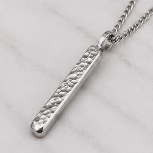 Könnte beinhalten: Eine silberfarbene Metall-Anhänger-Halskette mit einem langen, schlanken Stabdesign. Die Vorderseite des Stabes hat eine ausgeprägte, gehämmert texturierte Oberfläche, während die Seiten und die Rückseite glatt und poliert sind. Sie hängt an einer passenden Metallkette.
