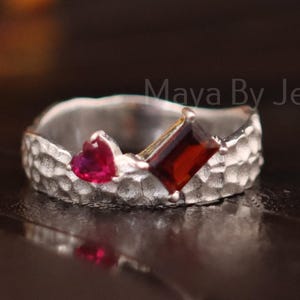 May include: A silver ring with a hammered texture, featuring a heart-shaped red gemstone and a square-cut red gemstone. The ring is from Mava By Jewelry.