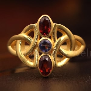 May include: A gold-colored ring featuring a Celtic knot design. The ring has three oval-shaped gemstones: two red and one blue. The ring is likely made of gold and is a piece of jewelry.