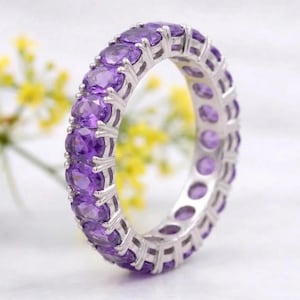 Op de afbeelding: Een zilveren eternity-ring met een doorlopende rij ovaal geslepen paarse amethist edelstenen in een klauwzetting. De metalen ring heeft een gepolijste afwerking en de paarse stenen zijn gelijkmatig rondom de gehele omtrek geplaatst.