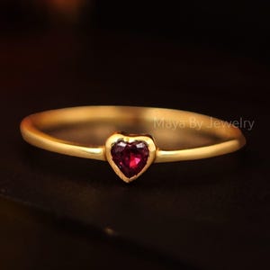 May include: A slender gold ring with a heart-shaped, deep red gemstone. The ring has a fine band and a simple, elegant design. The gemstone is set in a bezel setting, and the ring is shown against a dark background.