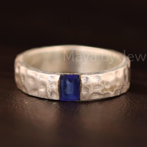 May include: A silver ring with a hammered texture and a rectangular, deep blue gemstone. The ring is a band style, with the gemstone set in the centre. The jewellery is displayed on a dark surface.