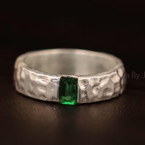 May include: A silver ring with a textured band and a rectangular, emerald-green gemstone. The ring is set against a dark background, highlighting the contrast between the metal and the vibrant stone. The ring is a piece of jewellery.