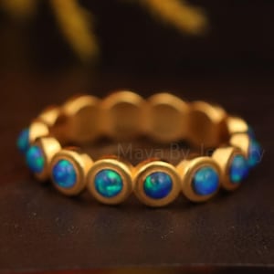 Könnte beinhalten: Ein goldfarbener Ring mit einer Reihe runder, blauer Opalsteine, die um das Band herum angeordnet sind. Die Opale haben eine Mischung aus blauen und grünen Farbtönen. Der Ring wird auf einer dunkelbraunen Oberfläche präsentiert.