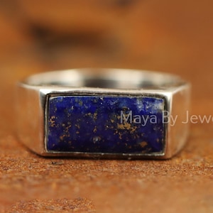 Op de afbeelding: Een zilveren ring met een rechthoekige, diepblauwe lapis lazuli steen. De steen heeft gouden vlekken en is gezet in een eenvoudige band. De ring is gemarkeerd met "Maya By Jewelry".