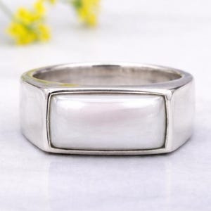 May include: A silver ring featuring a rectangular, pearlescent central stone. The ring has a modern, minimalist design with a polished finish. The background includes soft, blurred yellow and white flowers, adding a touch of natural elegance.