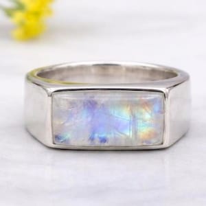 Op de afbeelding: Een zilveren ring met een rechthoekige, iriserende maansteen. De maansteen toont een scala aan kleuren, waaronder blauw, paars en geel. De ring ligt op een wit oppervlak, met gele bloemen op de achtergrond.