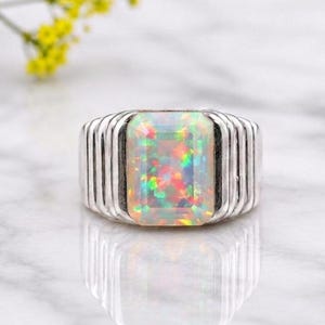 May include: A silver ring featuring a large, rectangular, faceted opal gemstone with a vibrant play of colour, including flashes of red, green, and yellow. The band is constructed with a series of vertical, parallel ridges on both sides.