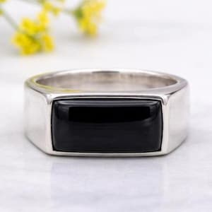 Peut inclure: Bague en argent avec une pierre d'onyx noire rectangulaire. La bague a un design simple et moderne, avec une finition polie. La pierre noire est sertie dans un sertissage à lunette. La bague est présentée sur une surface blanche.