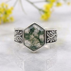 May include: A silver ring featuring a hexagonal moss agate gemstone with green dendritic inclusions. The band is crafted with a detailed embossed leaf pattern on both sides, framing the central stone in a polished metal setting.