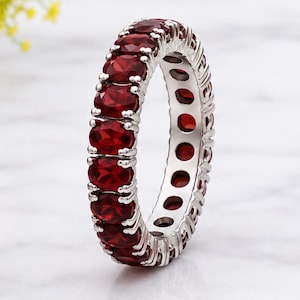 Puede incluir: Un anillo de eternidad que presenta una hilera continua de piedras preciosas de granate rojo oscuro de corte ovalado, engastadas en una banda de metal de tono plateado. Las piedras están dispuestas en un engaste de garras, creando un diseño circular.