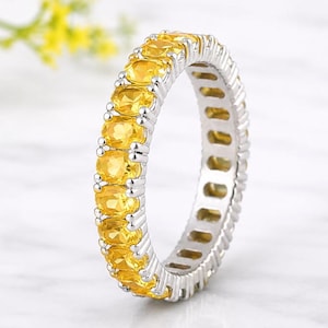May include: A silver-toned eternity band ring featuring a continuous row of oval-cut yellow gemstones set in a prong setting. The metal band has a polished finish and the stones are arranged in a uniform pattern around the entire circumference.